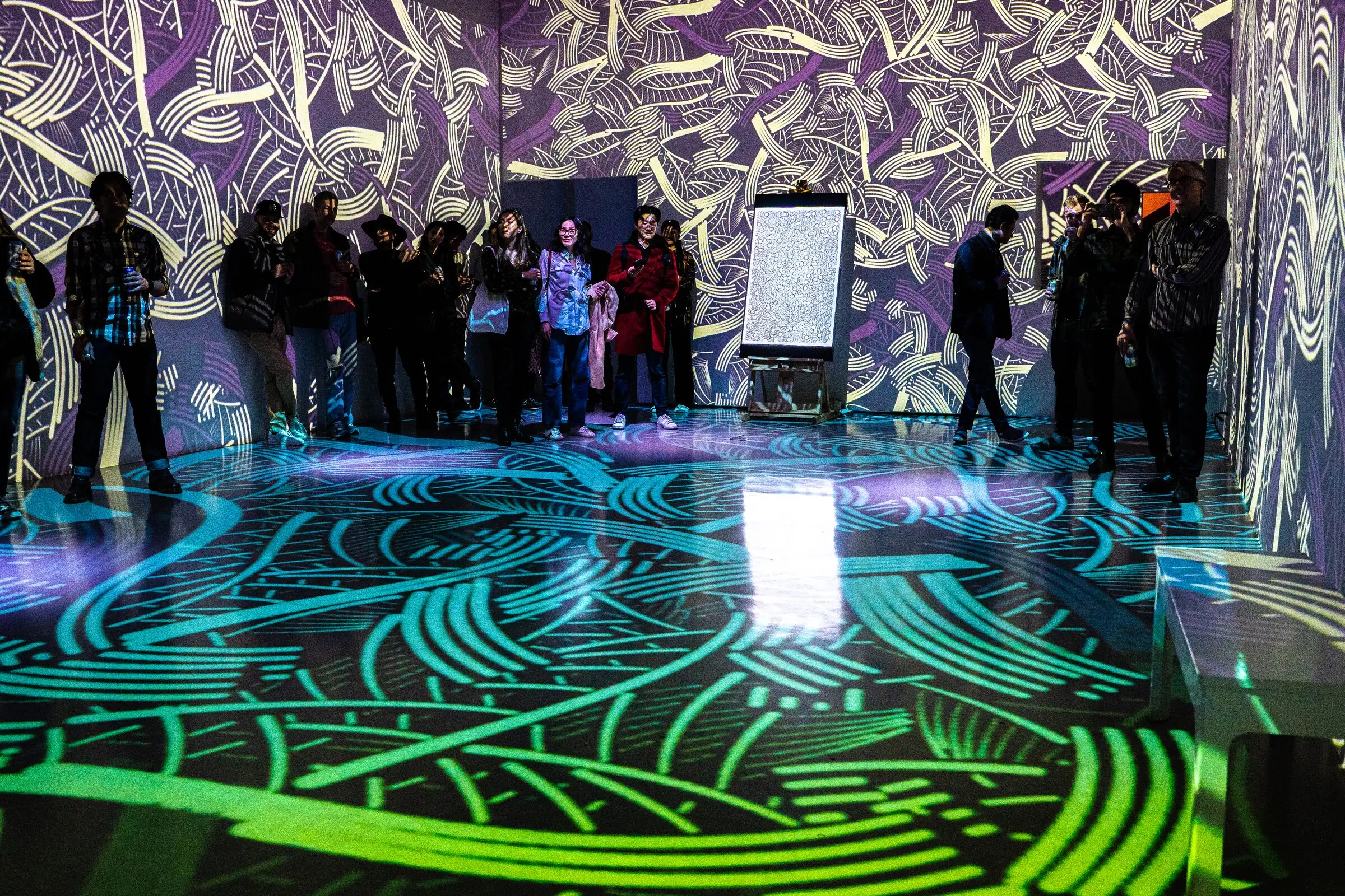 A crowd in a room with projected patterns on walls and floor during the INTRICADA event, Lume Studios, New York.